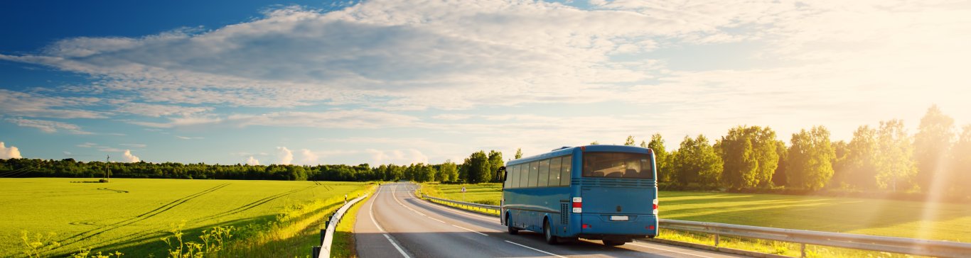 Ein blauer Bus fährt eine Landstraße entlang. Ringsum sind bewachsene Felder und ein blauer Himmel mit leichten Wolken zu sehen