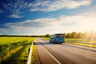 Ein blauer Bus fährt eine Landstraße entlang. Ringsum sind bewachsene Felder und ein blauer Himmel mit leichten Wolken zu sehen