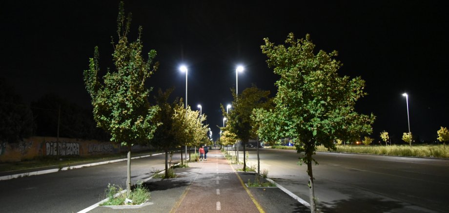 Breite Straße mit Rad-/Fußweg, welcher mit kleinen Bäumen gesäumt und mit Straßenlampen beleuchtet ist. In der Ferne sind zwei Fußgänger zu erkennen.