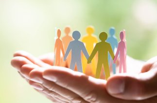 Zwei zur Schale geformte Hände auf denen bunte Papierfiguren mit menschlichen Umrissen in verschiedenen Farben einen Kreis bilden. Der grüne Hintergrund ist verschwommen.