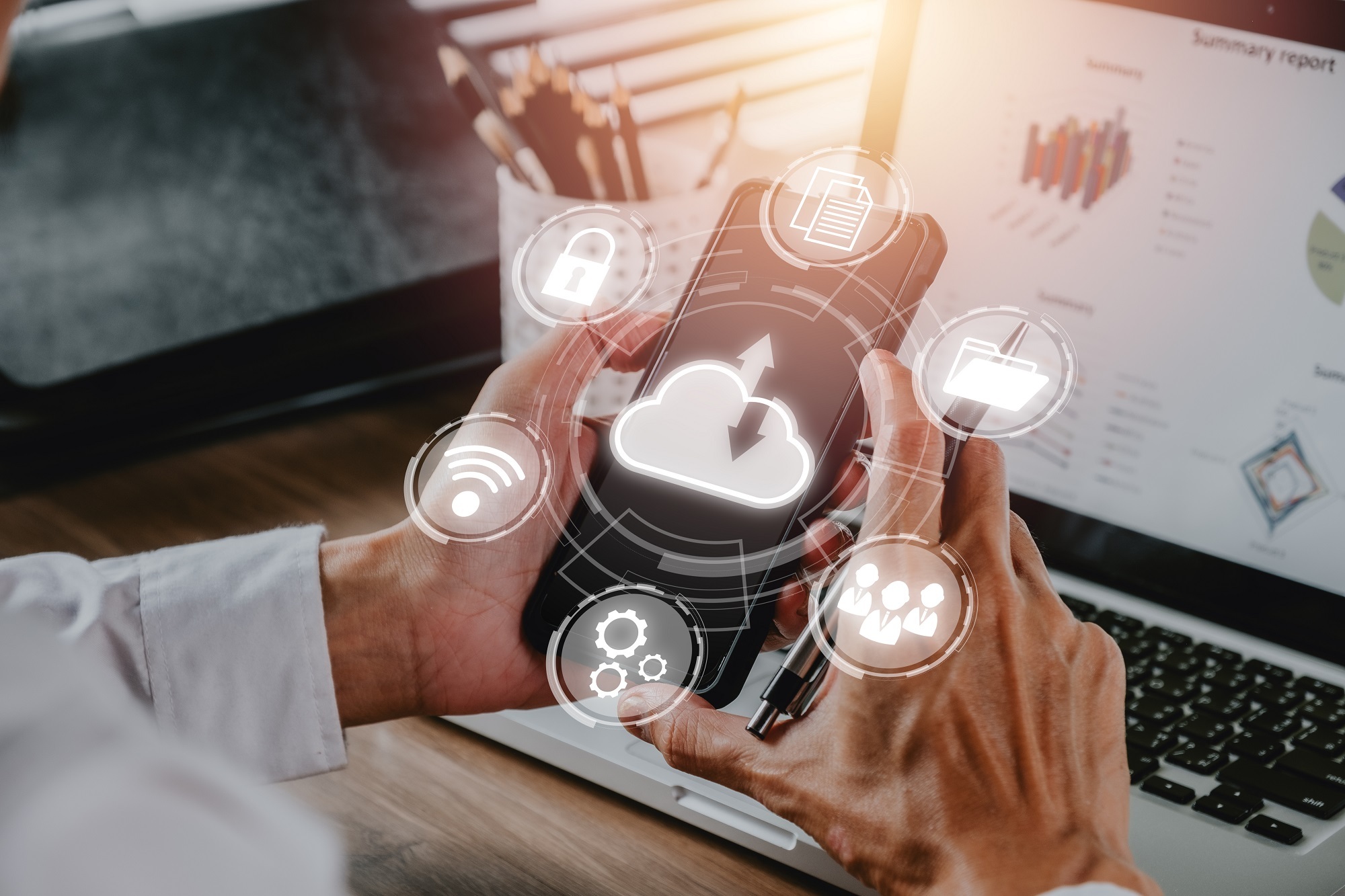 Männliche Hände halten ein Handy. Im Hintergrund ist ein Laptopbildschirm zu sehen. Im Vordergrund sind weiße Symbole abgebildet, welchen den up- und download in einer Cloud symbolisieren.