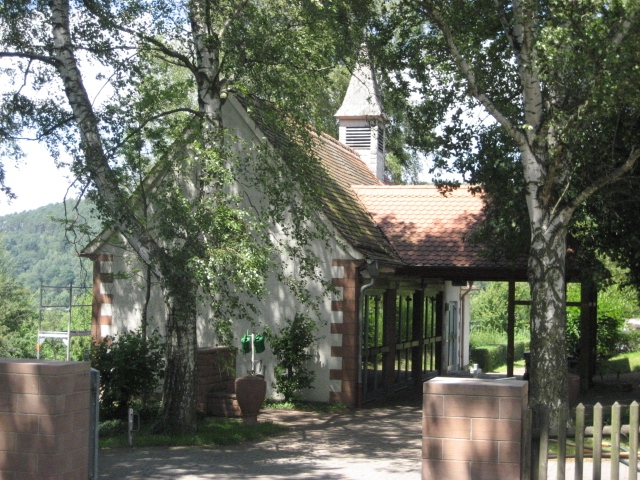 Trauerhalle am Friedhof im Ortsteil Eidengesäß, umgeben von Bäumen. Im Vordergrund ist der Eingangsbereich zum Friedhof zu erkennen.