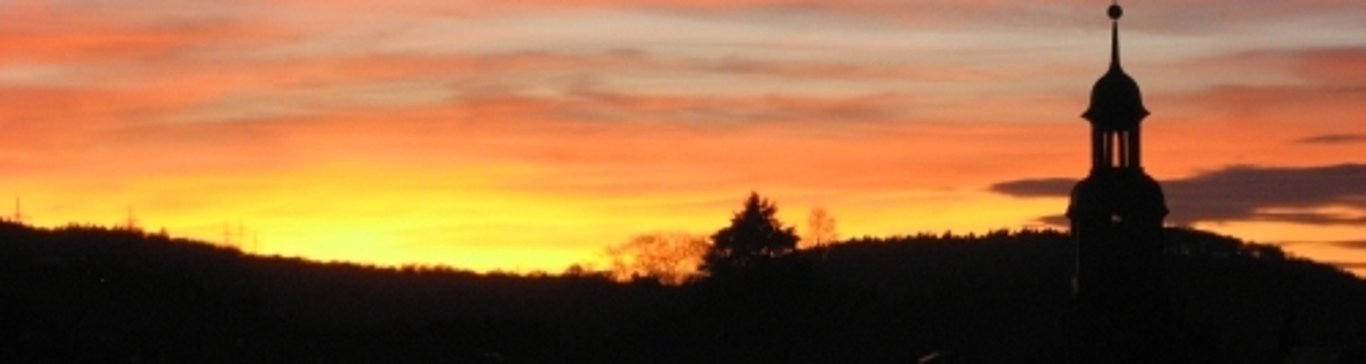 Sonnenuntergang mit orange gefärbtem Himmel. Eine schwarze Silouette von Bäumen und Hügeln ist zu erkennen. Ebenso die schwarzen Umrisse eines kleinen Glockenturmes im rechten Bildbereich.