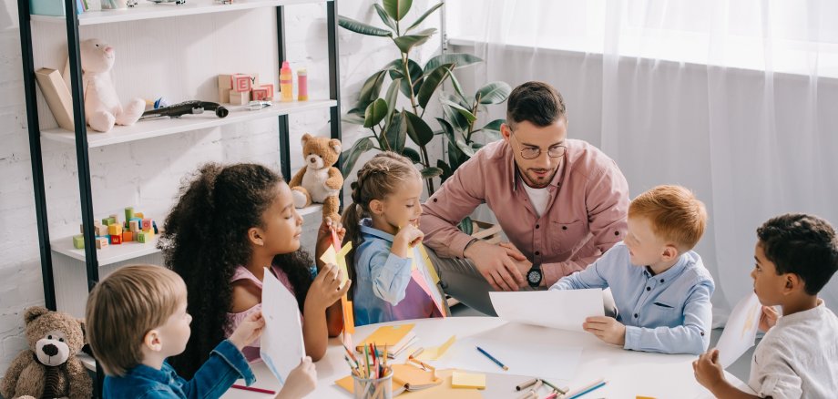 teacher and interracial preschoolers at table with paints and pa