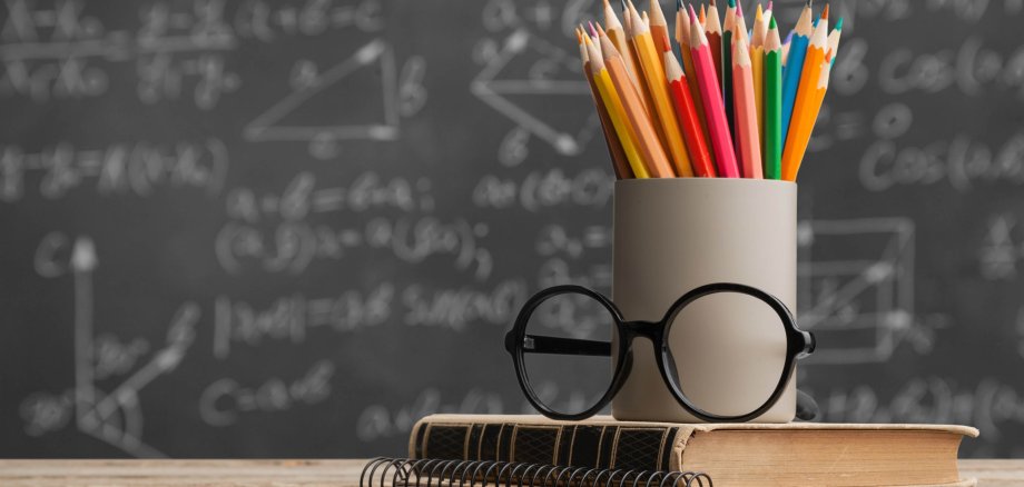 Farbige Stifte in einem Becher, Buch und Brille stehen vor einer Tafel, die mit mathematischen Formeln beschriftet ist
