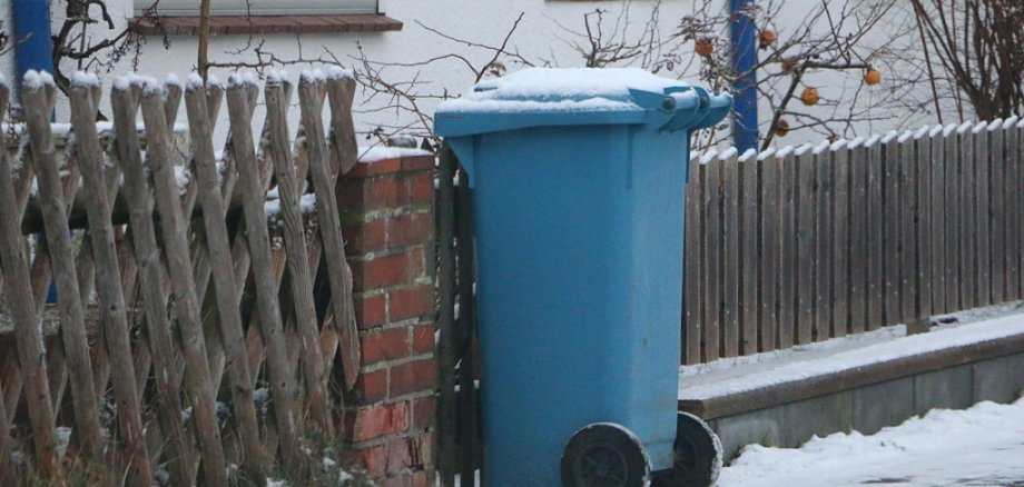 blue plastic recycling bin for paper waste stands at the roadside, covered in snow, ready for collection