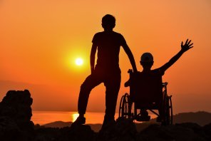 Eine person im Rollstuhl und eine stehende Person blicken gemeinsam in den Sonnenuntergang