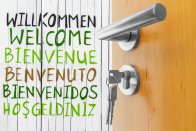 Hinter eine mit Schlüssel geöffneten Tür heißt der Schriftzug Willkommen in Verschiedenen Sprachen willkommen