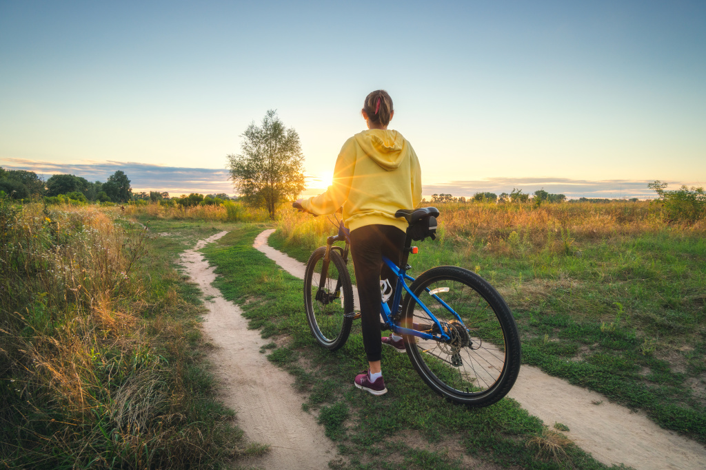 Woman is riding a mountain bike in cross country road at sunset in summer. Colorful landscape with sporty girl in yellow hoodie, bicycle, field, dirt road, green grass, trees, sky. Sport and travel Woman is riding a mountain bike in cross country road at sunset in summer. Colorful landscape with sporty girl in yellow hoodie, bicycle, field, dirt road, green grass, trees, sky. Sport and travel