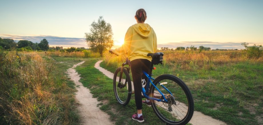 Woman is riding a mountain bike in cross country road at sunset in summer. Colorful landscape with sporty girl in yellow hoodie, bicycle, field, dirt road, green grass, trees, sky. Sport and travel